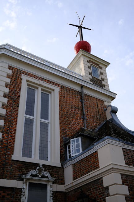 A close-up view of the upper portion of a historic brick building showing architectural details such as large, vertical sash windows with white frames and stained glass elements, decorative stonework above a smaller window, and a sloped slate roof. The building features a tower-like structure topped with a red spherical ornament and a metal weather vane with elongated arrow-shaped blades. The sky in the background is clear and light blue, indicating daytime. The scene emphasizes the traditional brick finish and the detailed craftsmanship of the facade, aligning with themes of property maintenance and private property management, which may relate to waste management or rubbish removal services in historic or private buildings. The image underscores the importance of building upkeep and the potential for on-site clearance of waste materials associated with property improvements or renovations, as handled by independent waste disposal providers like Waste Disposal Richmond upon Thames. The overall setting appears to be the exterior of a well-maintained historic building, with no visible waste or rubbish present in the image itself.