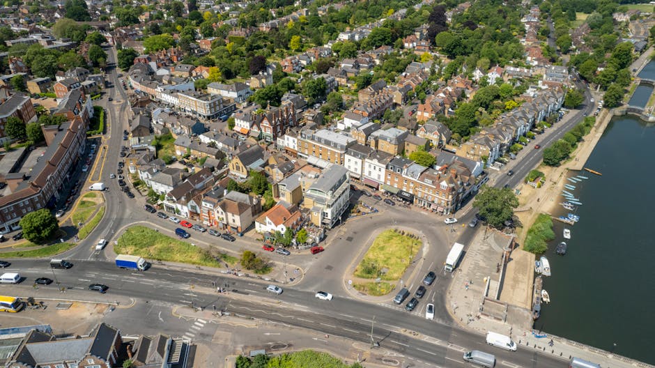 An aerial view of a bustling town centre near a river, featuring a mix of Victorian and modern brick buildings in shades of red, brown, and white, with flat and pitched roofs. The foreground shows a wide intersection with multiple lanes, parked cars, and a few moving vehicles, surrounding a small roundabout with patches of grass. To the right, a paved promenade runs along the riverbank, where several boats are moored, and a paved walkway lined with trees provides access to the water. The area includes parking spaces and a small open area with a few benches and trees, indicating a community space or waiting zone. The background reveals a densely populated residential district with narrow streets, tree-lined avenues, and closely packed houses with rooftops in various shades of slate and terracotta. This urban landscape emphasizes the typical elements of a town centre that may require private waste management or occasional rubbish clearance, as part of maintaining the area's cleanliness and appeal. Waste disposal or rubbish removal services in Richmond often involve on-site collection or private clearance to manage the waste generated in such busy local environments, aligning with the context of alternative waste handling methods for commercial and residential areas.