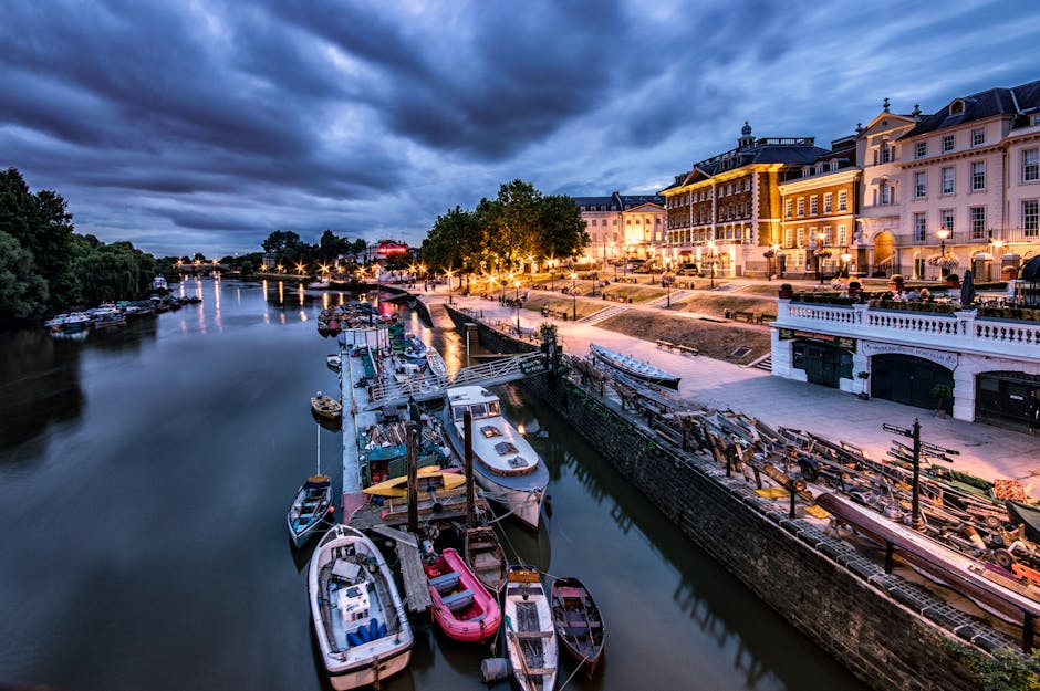A wide view of a riverbank at dusk showing a collection of boats docked along a wooden pier, with some boats covered in tarps and others open revealing seating and equipment. The river's calm waters reflect the evening sky and the lights from the surrounding buildings. On the right side of the image, a row of multi-story historic-style buildings with large windows, ornate facades, and exterior lighting line a paved promenade. Several street lamps illuminate the walkway, which is partially landscaped with small trees and benches. In the background, the river continues toward the horizon with more boats visible, and a dark, cloud-filled sky adds a dramatic atmosphere, suggesting a lively waterside area that may attract leisure activities, with potential relevance to private waste disposal services like those offered by waste disposal Richmond upon Thames.